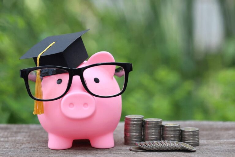 A pink piggy bank with a graduation cap placed on top of it and a pair of glasses placed over its nose, set beside stacks of coins