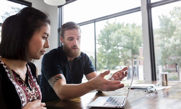 A man and a woman sitting side by side, looking at something on the laptop placed on a table in front of them