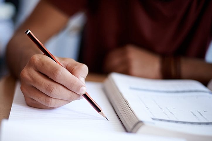 A closeup of a hand holding a pencil over a piece of notebook paper with an opened book beside their hand
