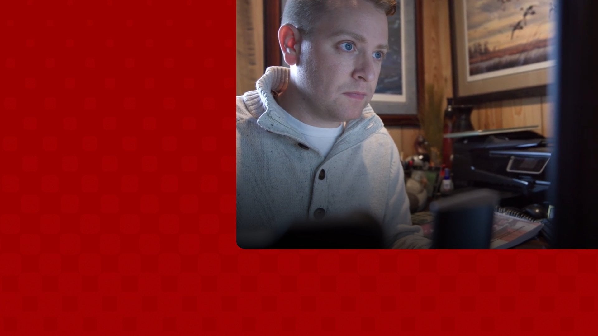 Man in grey sweater looking at computer screen with framed art in background.