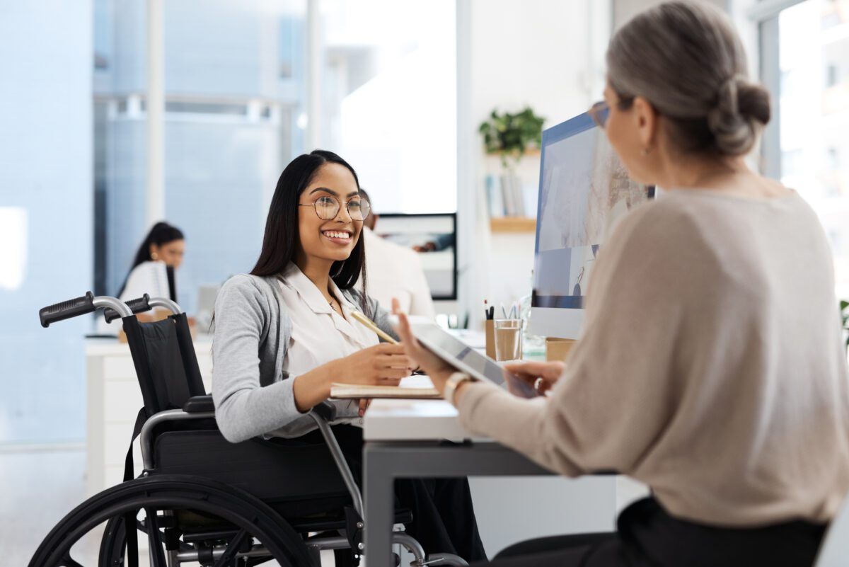 women at desk smiling, sitting in a wheelchair, looking at another woman across from her