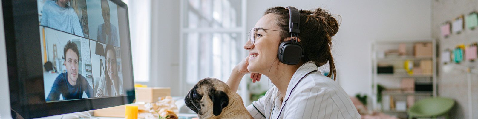Woman wearing headphones and holding a pug talks to colleagues in a video conference from an office setting