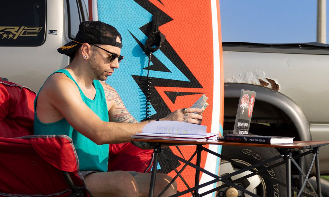Joey sitting next to his van and surfboard with a foldout table and his laptop