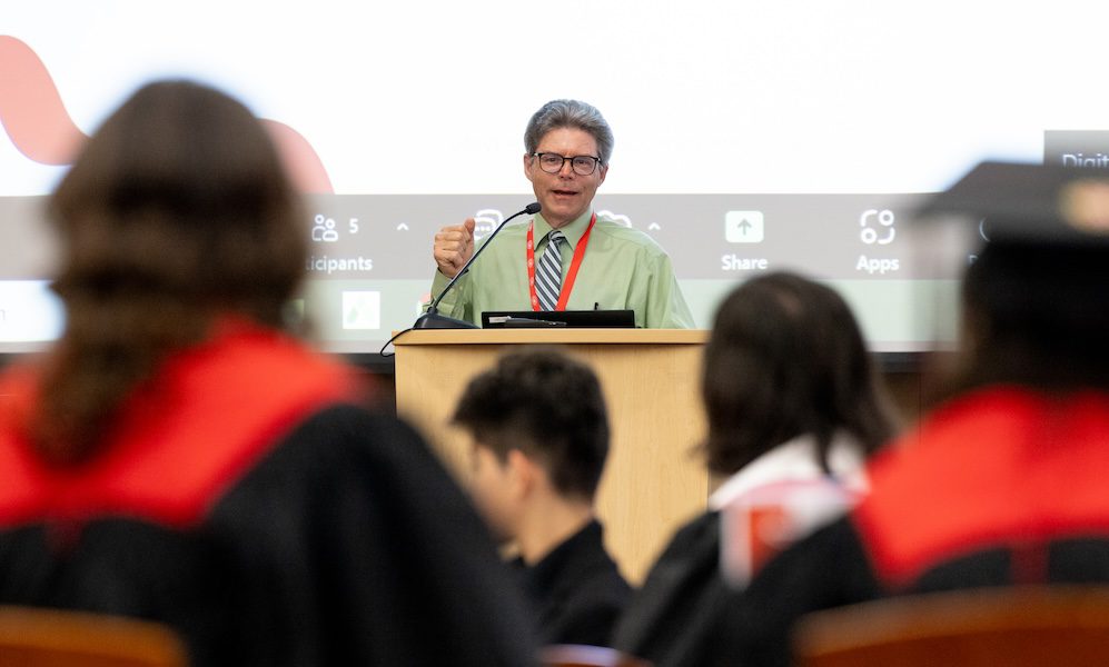 Jeff Russell speaks behind a podium during the UW–Madison Online graduation ceremony. He is wearing a long-sleeve green shirt, glasses and a red lanyard and has short gray hair. Audience members in the foreground are slightly blurred; several are wearing graduation gowns.