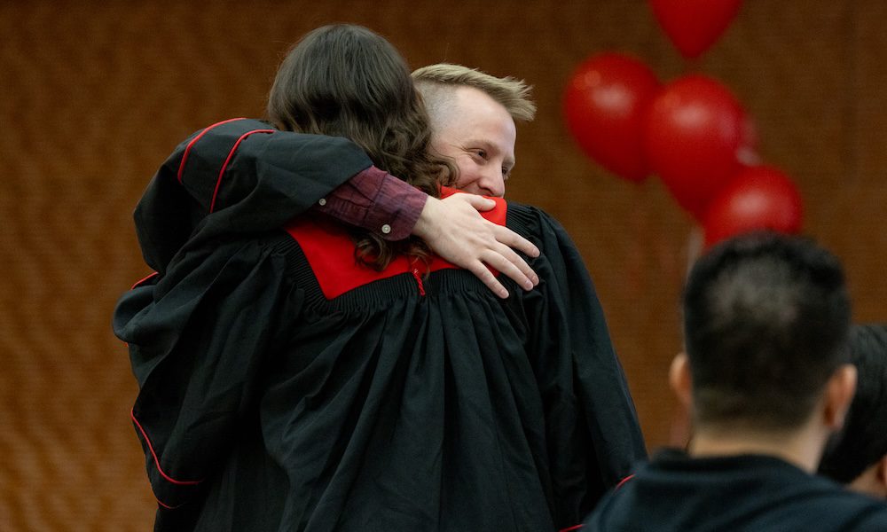 UW–Madison Online graduates Manny Avila and John Gloeckler share a hug. Both are wearing graduation gowns. Manny's back faces the camera. John's hand and smiling face are visibile over Manny's shoulder.