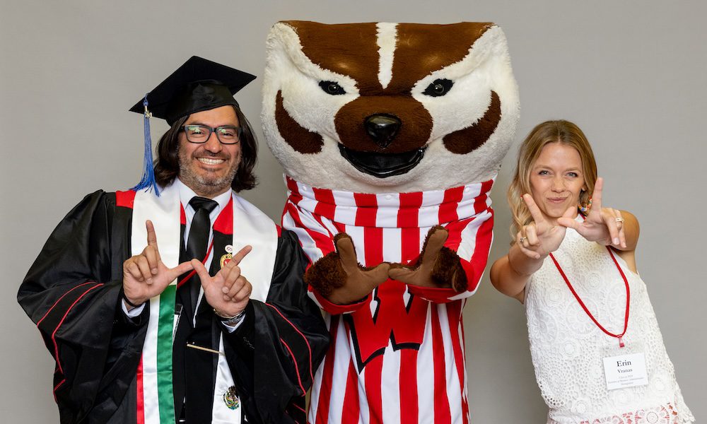 UW–Madison Online graduates Manny Avila and Erin Vranas stand on either side of Bucky Badger flashing the "W" with their fingers. Manny, on left, has dark hair and glasses and is wearing his graduation cap and gown. Erin, on right, is wearing a white sleeveless top and has long blonde hair.