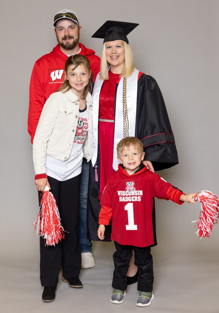 Kristi Jorgensen, UW–Madison Online graudate, poses against a plain backdrop with her family. Kristi is wearing her cap and gown and has long blonde hair. She is accompanied by her husband, daughter and young son. Both of her children hold red and white pom-poms.