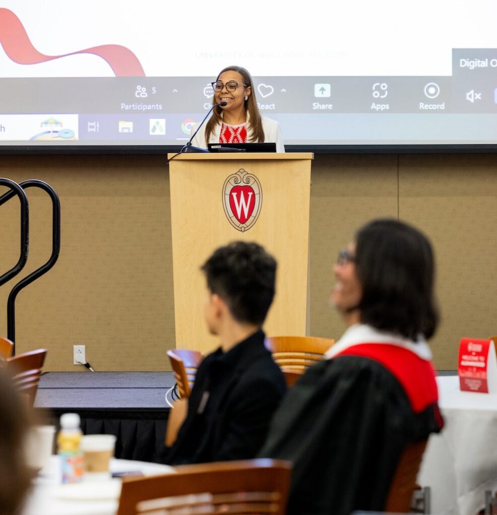 Dr. Maya Evans addresses the audience of the UW–Madison Online graduation. Audience members in the foreground are blurred slightly. Maya stands behind a lectern with the UW–Madison crest on the front. She has long dark hair and wears glasses, a red pattern top and a white suit jacket.