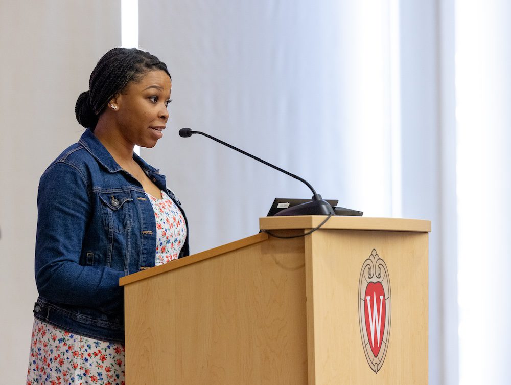 Karen Thomas, viewed from the side, stands at a lectern with the UW–Madison crest on the front and speaks into a microphone. She is wearing a flower print dress and jean jacket. She has dark hair pulled back into a bun.
