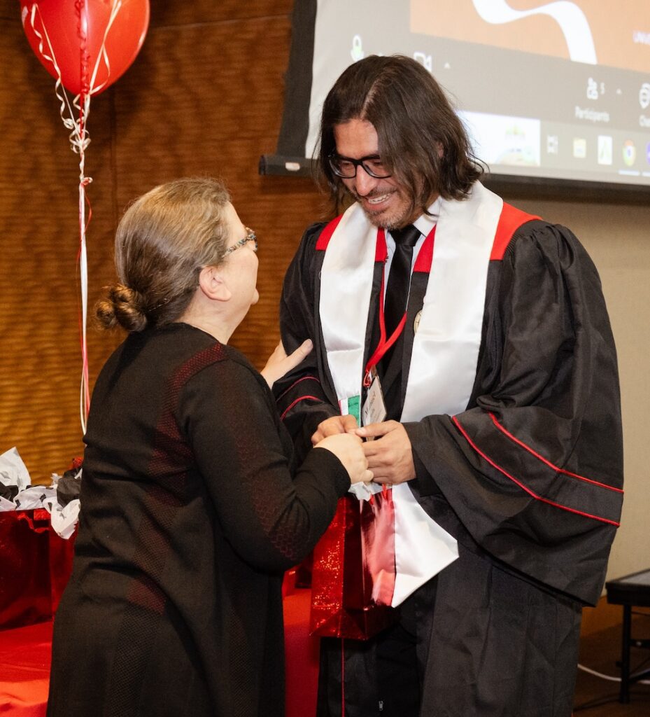 UW–Madison Online advisor Leanne Johnson presents grad Manny Avila with a red gift bag. They look at one another and smile as they hold hands. Leanne has dark blonde hair pulled back and is wearing a black top and glasses faced mostly away from the camera. Manny wears his graduation robe. Red balloons are in the background.