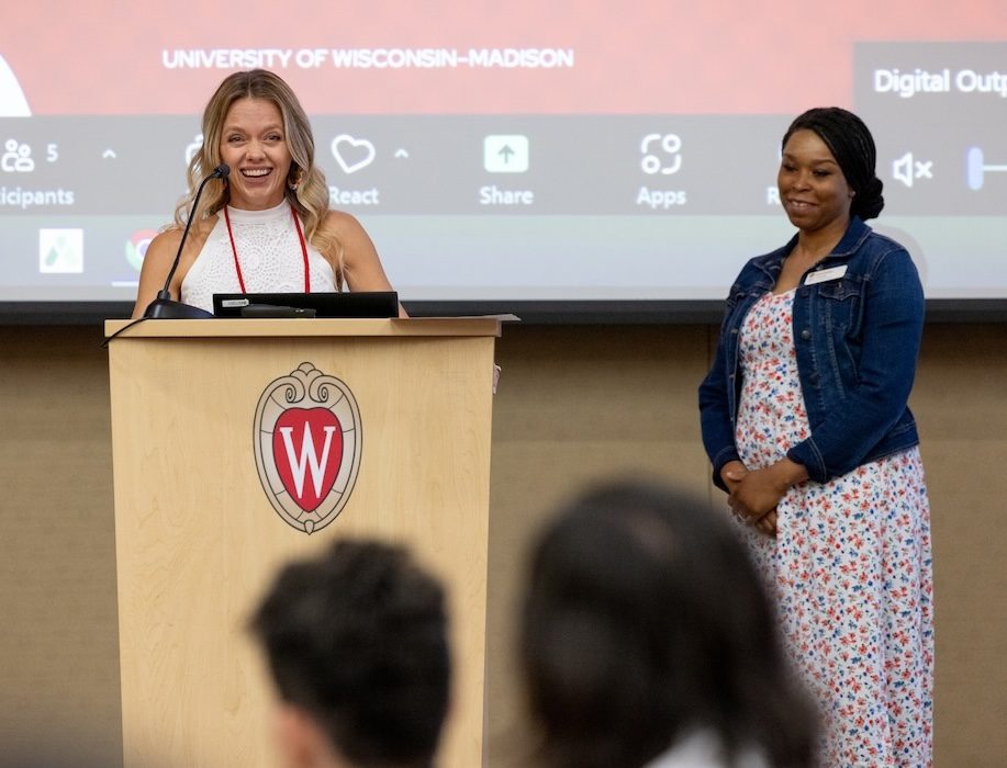 Erin Varnas faces the crowd at UW–Madison Online's graduation. She is smiling while she speaks, has long blonde hair and is wearing a sleeveless white top. Karen Thomas stands near her on stage, looking on as Erin makes her comments. Karen is wearing a floral white dress, jean jacket and has dark hair pulled back in a bun. 