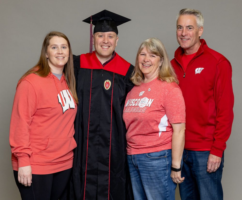 UW–Madison Online graduate John Gloeckler poses in his cap and gown with his parents and wife. John has blonde hair. His parents and wife are wearing various red UW shirts and are smiling. 