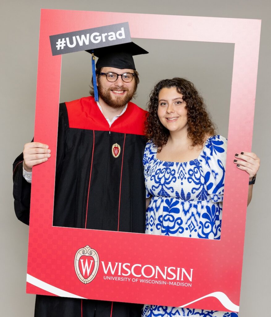 UW-Madison Online graduate stands in cap and gown next to his wife. Troy has medium long dark hair and wears dark glasses. His wife wears a blue and white floral dress and has long, dark curly hair. They are standing within a large red photo frame, which they hold out in front of them. The photo frame has the UW–Madison logo on the front along with the words #UWGrad 
