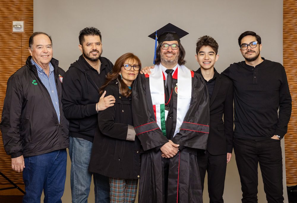 UW–Madison Online grad Manny Avila stands between five family members wearing his UW–Madison cap and gown. Family members range in age from his teenage son to older adults.